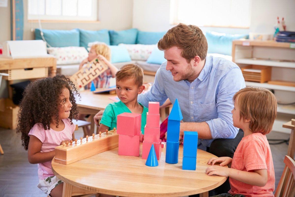Male Montessori teacher acting as a guide, interacting with children working on various sensorial materials like the number rods and geometric solids in a mixed-age classroom