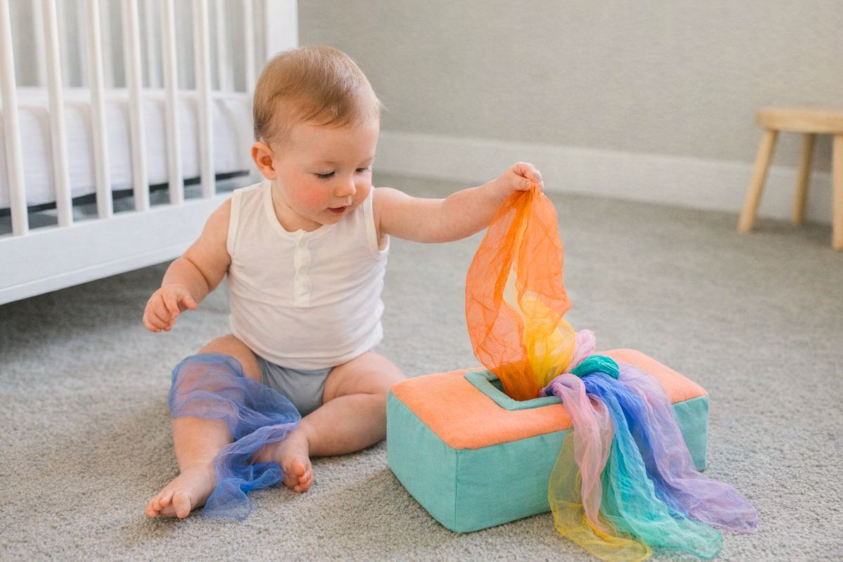6-month-old baby sitting and pulling soft colorful scarves out of a sensory tissue box toy