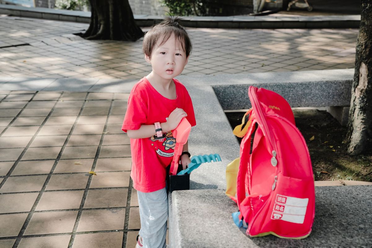 Small child standing next to a red school backpack on a sidewalk