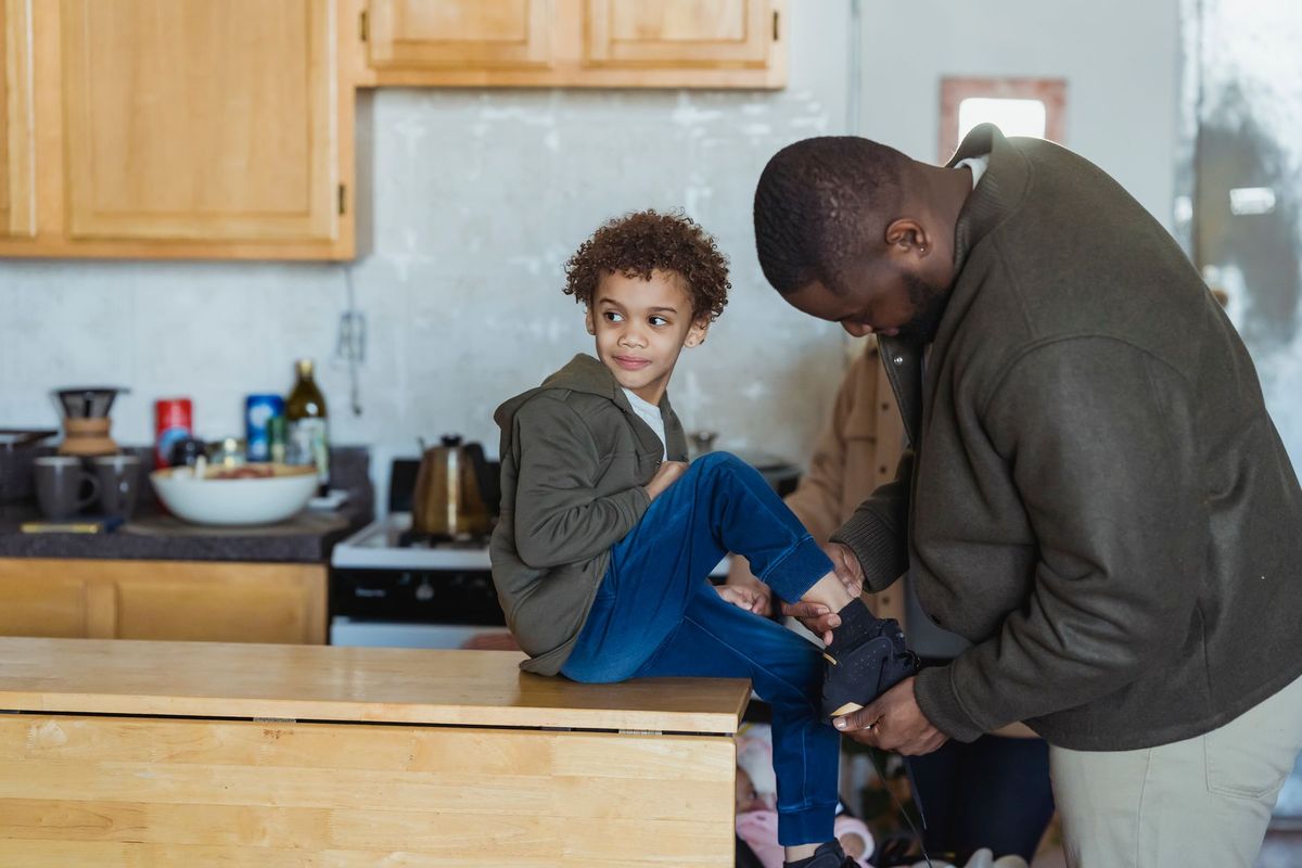 Father kneeling to help a young boy put on his shoes in the kitchen