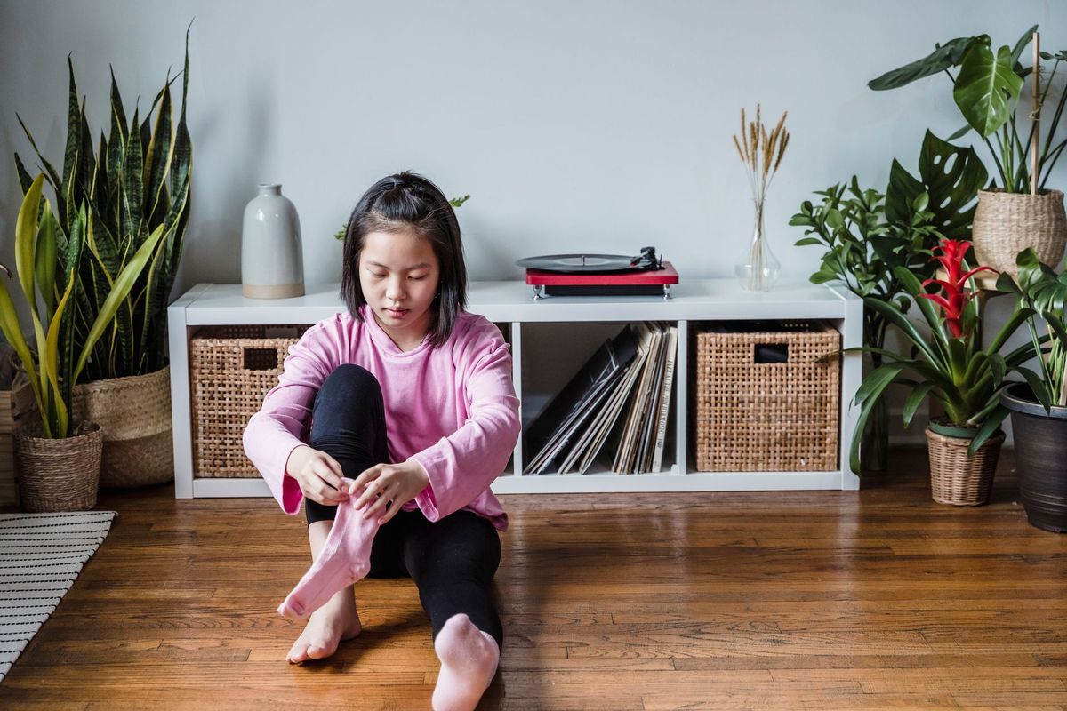 Young girl sitting on the floor putting on her socks by a low shelf