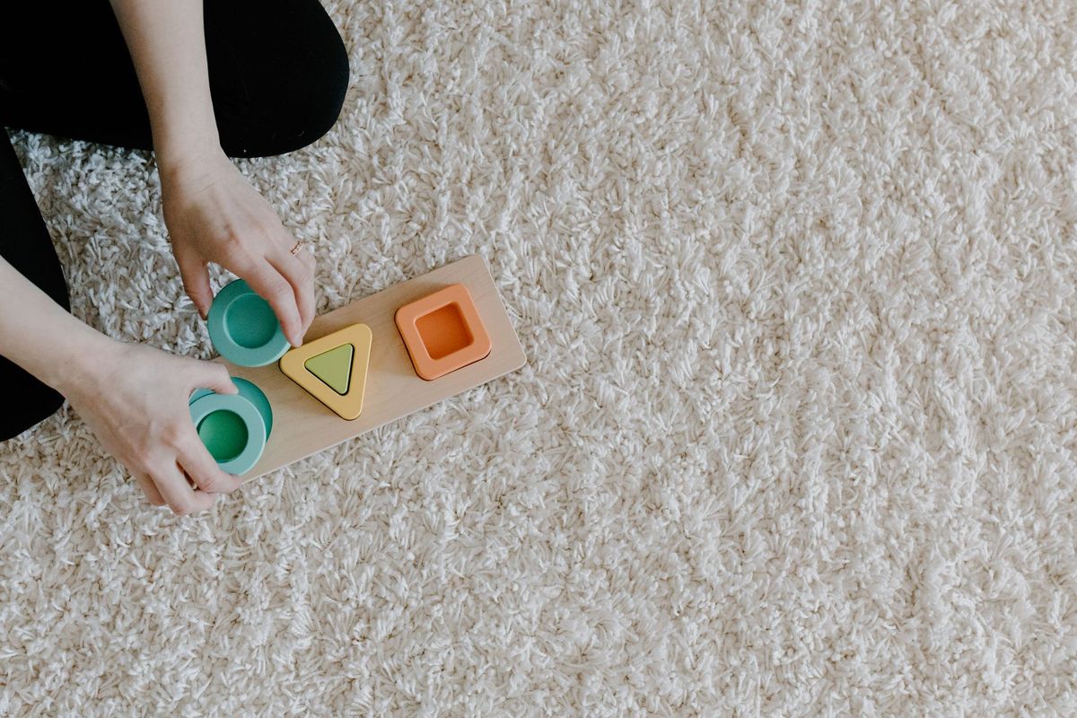 Child's hands placing chunky shapes into a wooden shape puzzle on a rug